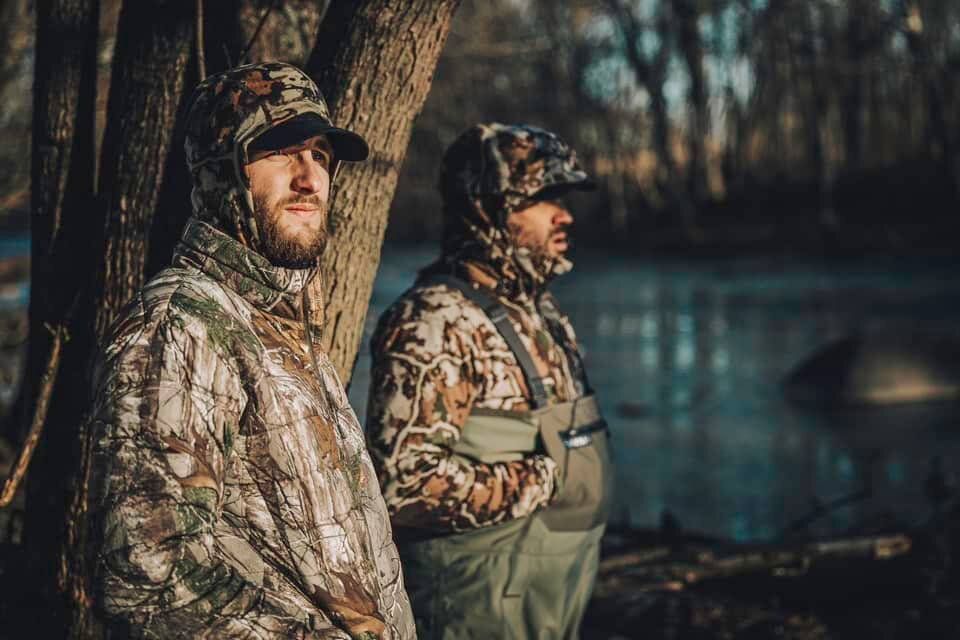 Two members of Hunt Moxie in camo standing by a tree. 