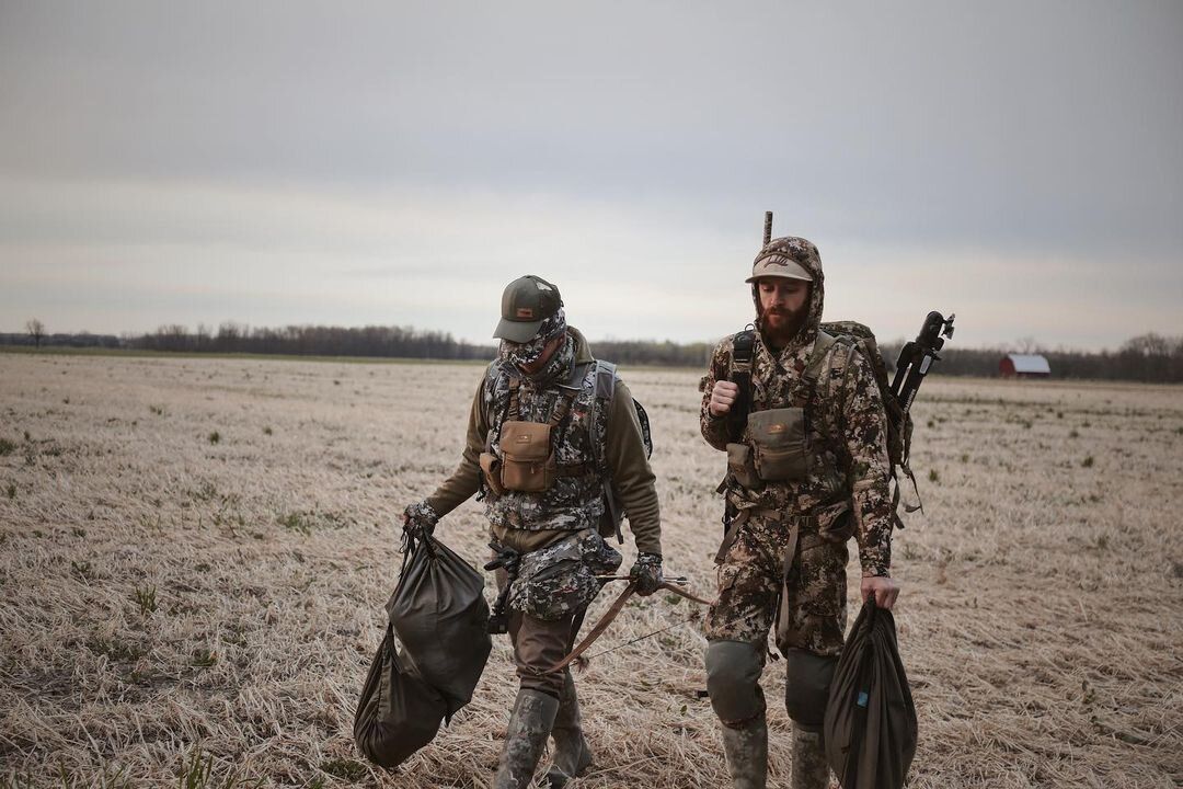 Two members of Whitetail Venatic wear camo and carrying hunting gear in a field. 