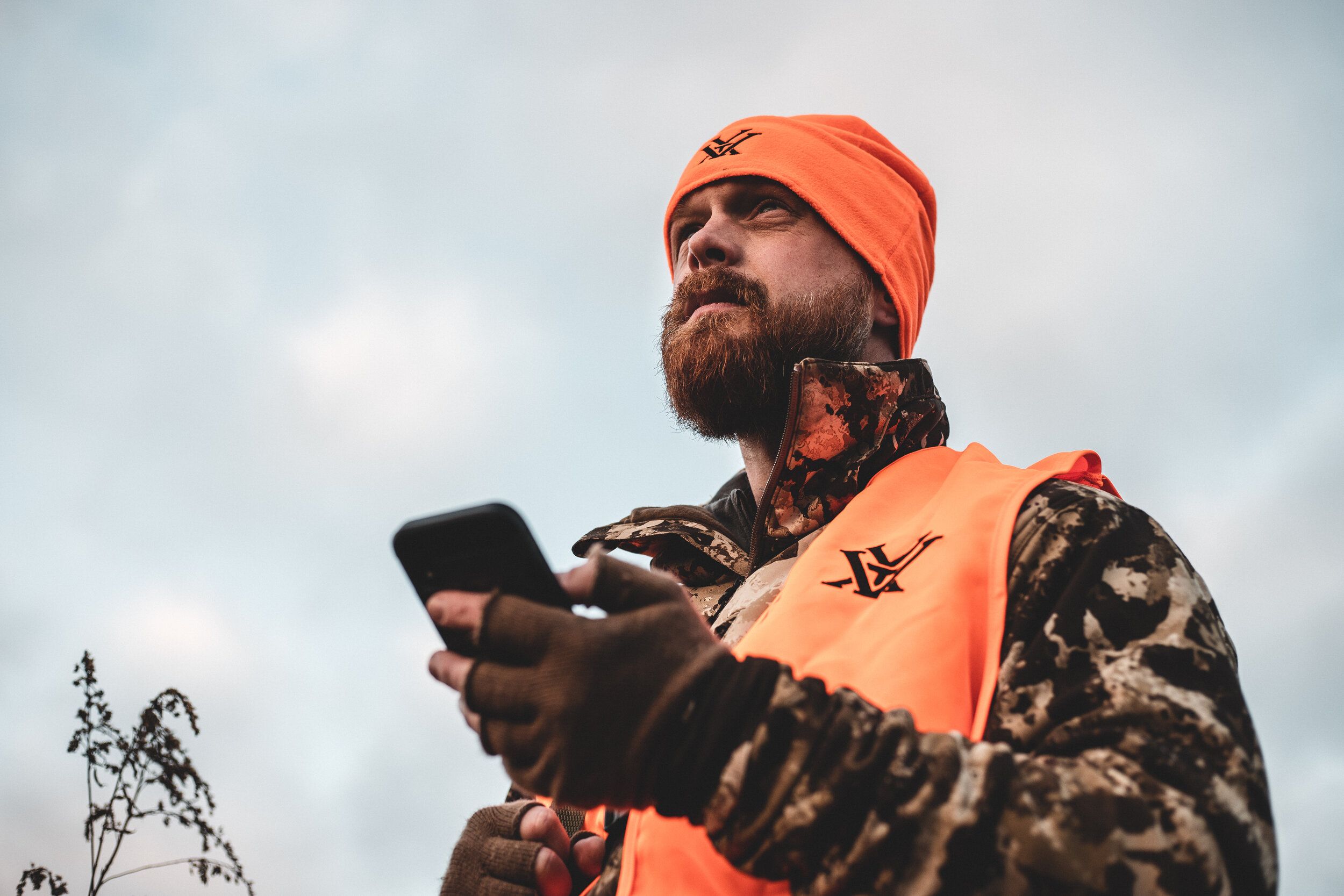 A hunter in blaze orange looks on while holding a cellphone, use HuntWise for late-season deer hunting concept. 