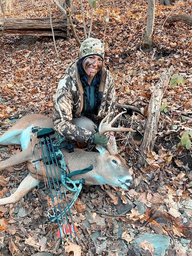 A female hunter sits on the ground with a deer kill and bow, using HuntWise concept. 