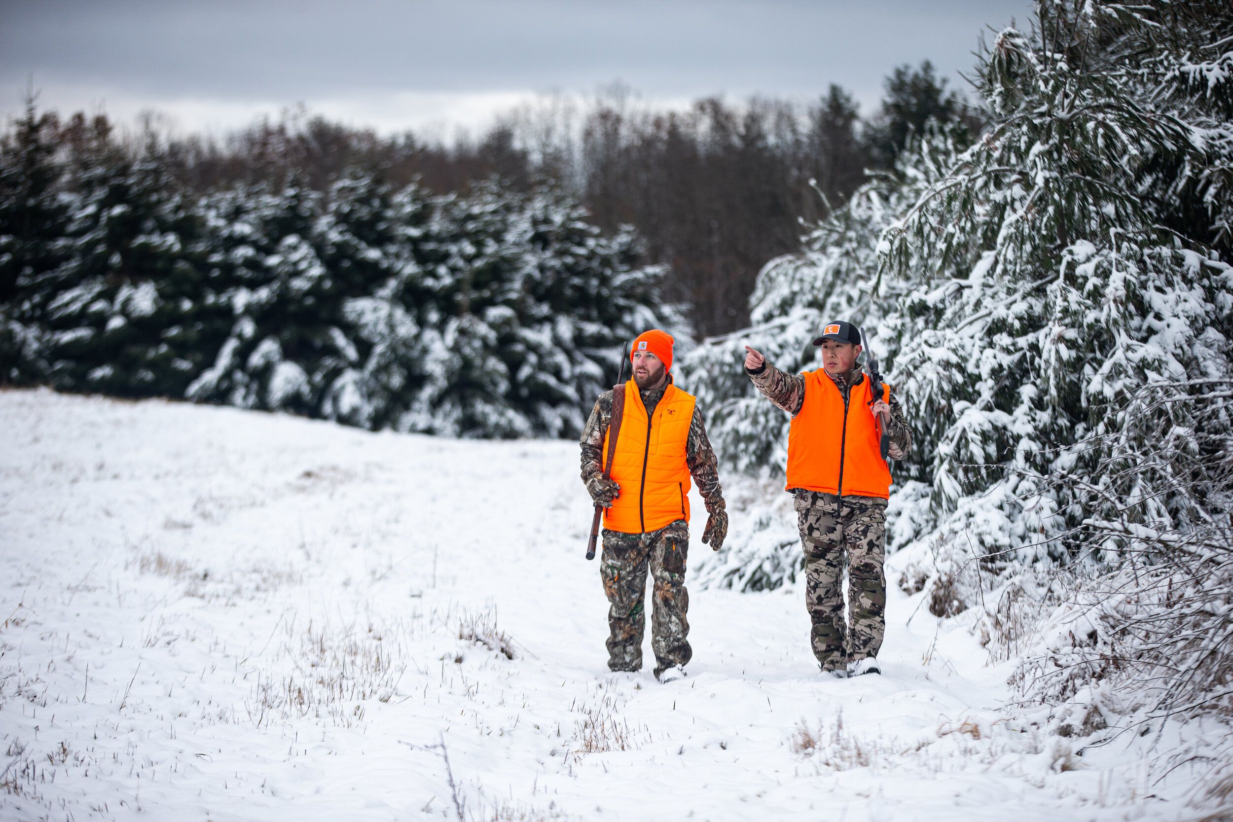 Two hunters walk in the snow near trees while wearing blaze orange, late-season whitetail hunting concept. 