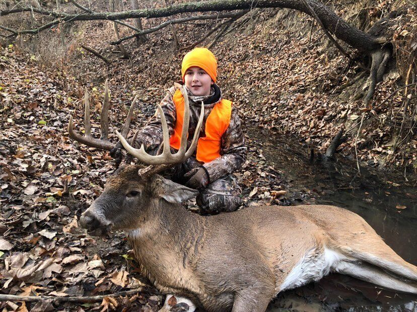 A boy wearing blaze orange holds up the antlers and head of a deer kill, hunting stories concept. 