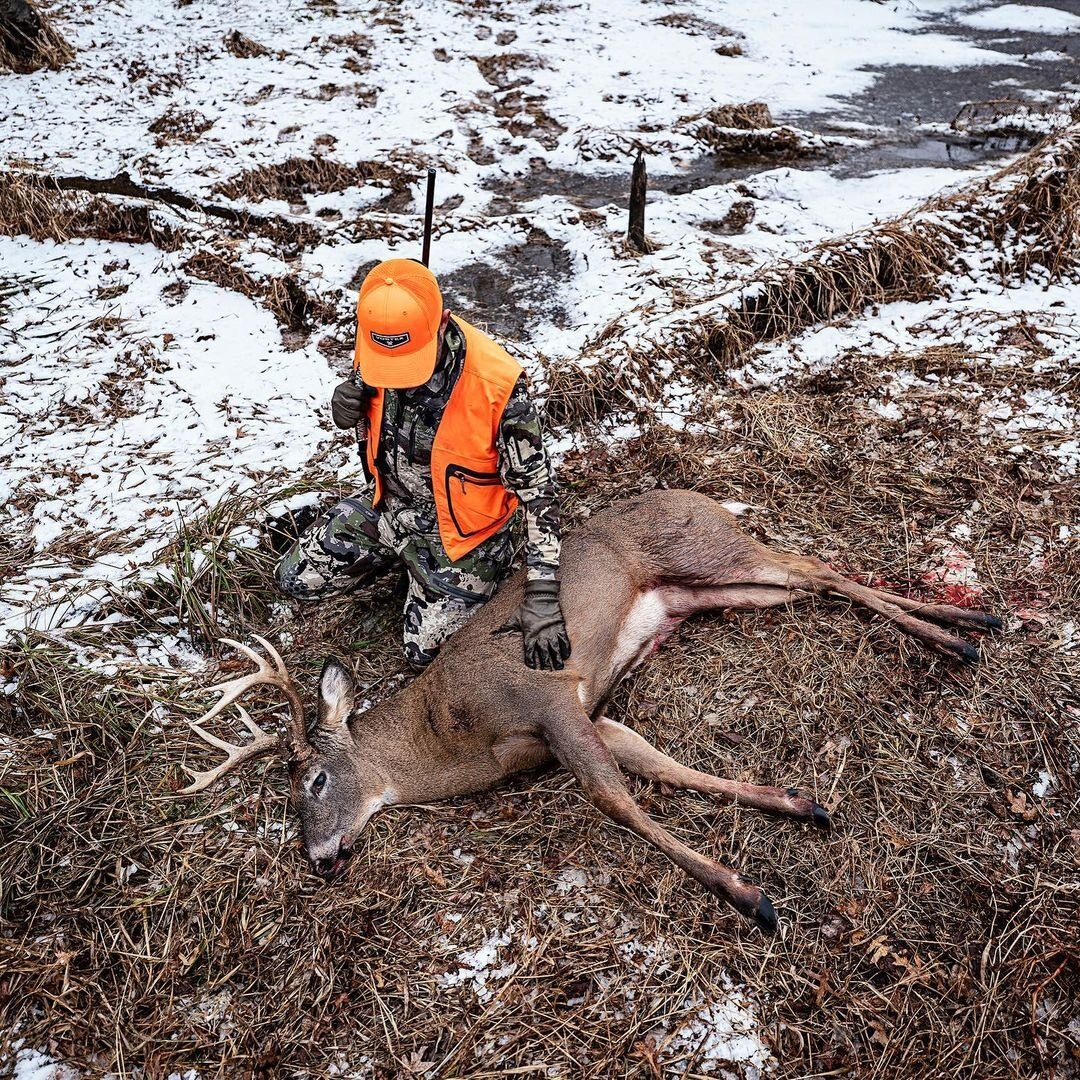 A hunter in blaze orange with a rifle kneels beside a deer kill, hunting stories concept. 