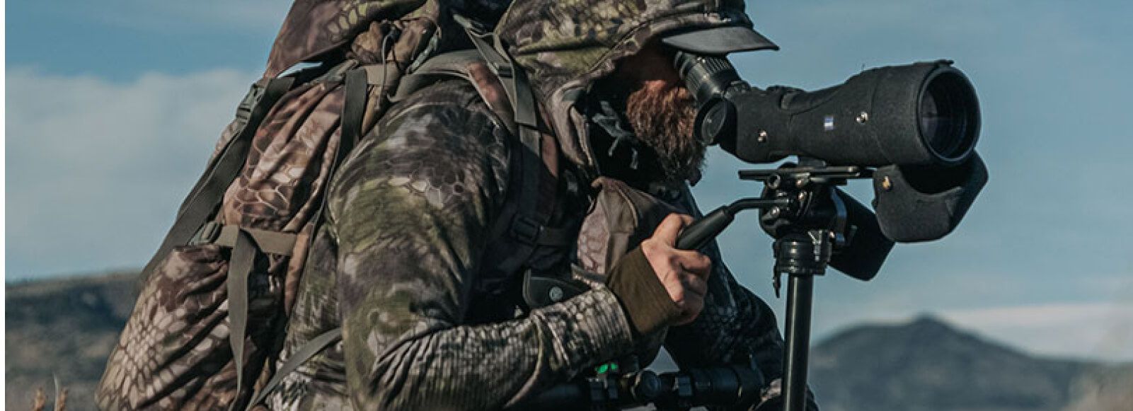 A hunter in camo with gear used a spotting scope. 