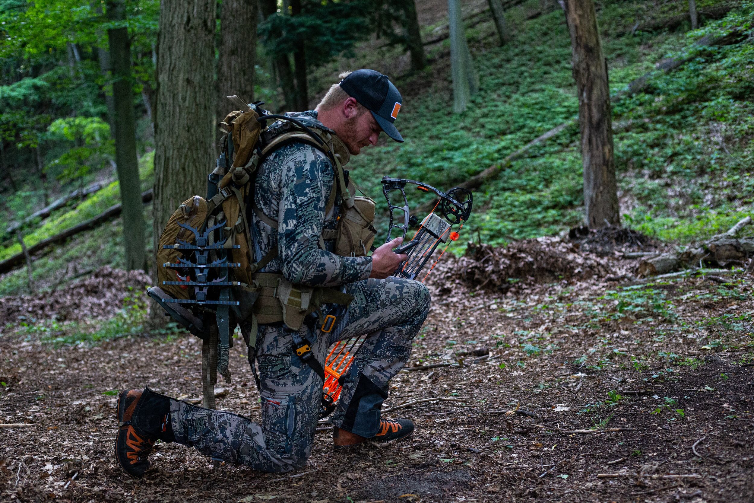 A hunter in camo kneeling on the ground in the woods looking at a phone, use HuntWise to find whitetail bucks. 